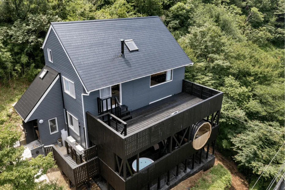 緑に囲まれた青い屋根の建物と黒いウッドデッキ(バレルサウナ・ジャグジー)の空撮